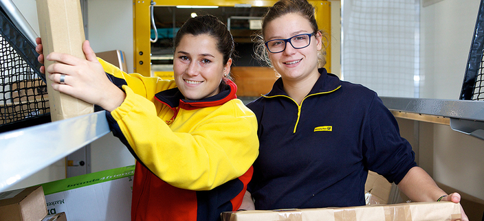 2 junge Frauen mit Post-Arbeitskleidung sortieren Pakete in ein Regal ein und lächeln 2 junge Frauen mit Post-Arbeitskleidung sortieren Pakete in ein Regal ein und lächeln