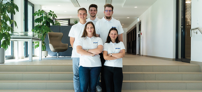 5 junge Menschen stehen mit weißen Firmenshirts in einem Gebäude vor drei breiten Stufen 5 junge Menschen stehen mit weißen Firmenshirts in einem Gebäude vor drei breiten Stufen