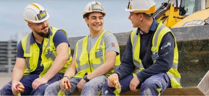 Drei Mitarbeitende von PORR in Arbeitskleidung in einer Baggerschaufel auf einer Baustelle sitzend Drei Mitarbeitende von PORR in Arbeitskleidung in einer Baggerschaufel auf einer Baustelle sitzend