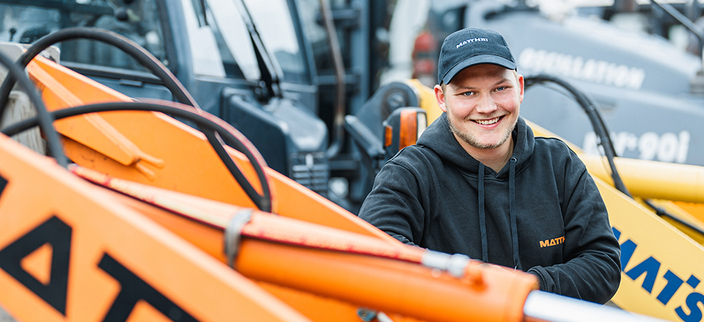 Junger Mensch in Kapuzenpulli und Basecap mit Firmenlogo steht zwischen Baumaschinen und lächelt in die Kamera Junger Mensch in Kapuzenpulli und Basecap mit Firmenlogo steht zwischen Baumaschinen und lächelt in die Kamera
