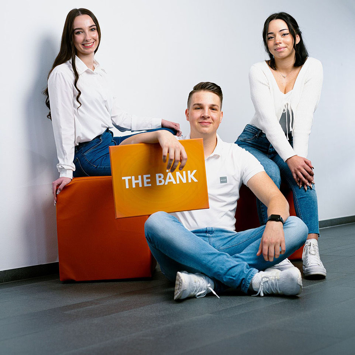 Personen sitzen zusammen, eine hält ein Schild hoch auf dem steht "THE BÄNK".