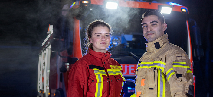 Zwei junge lächelnde Personen in Feuerwehrbekleidung vor dem Einsatzwagen Zwei junge lächelnde Personen in Feuerwehrbekleidung vor dem Einsatzwagen