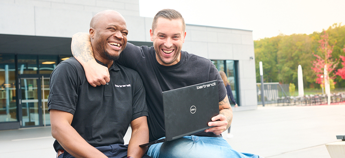 Zwei Personen sitzen auf einer Bank. Die eine Person hat einen Laptop mit der Aufschrift "bertrandt" auf dem Schoß und legt der anderen Person den Arm um den Hals. Beide Personen lachen. Zwei Personen sitzen auf einer Bank. Die eine Person hat einen Laptop mit der Aufschrift "bertrandt" auf dem Schoß und legt der anderen Person den Arm um den Hals. Beide Personen lachen.