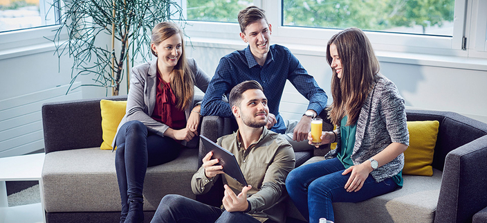 Drei junge Personen sitzen auf einem Sofa, eine Person sitzt vor dem Sofa und zeigt etwas auf einem Tablet