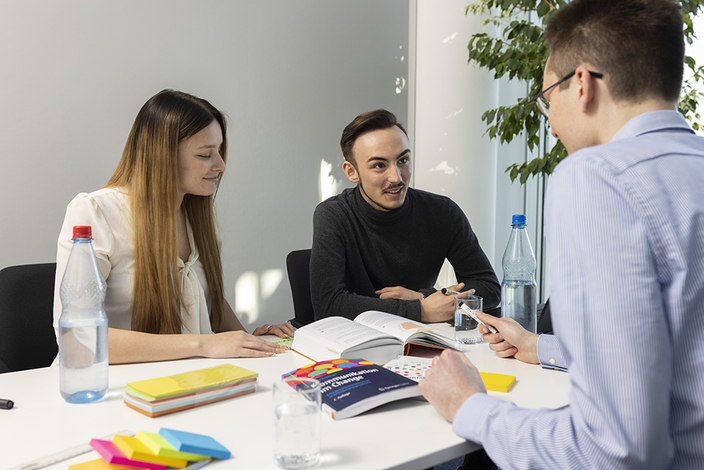 Drei Personen sitzen an einem Tisch und unterhalten sich. Auf dem Tisch liegen Bücher und weitere Arbeitsmaterialien.. Drei Personen sitzen an einem Tisch und unterhalten sich. Auf dem Tisch liegen Bücher und weitere Arbeitsmaterialien..
