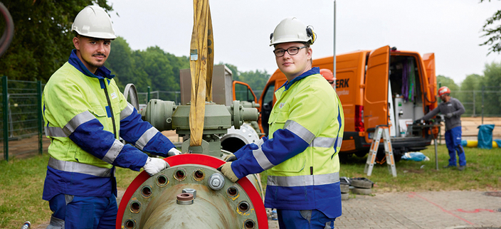 Zwei Mitarbeitende in Arbeitskleidung mit Helm, im Hintergrund ein orangefarbener Transporter Zwei Mitarbeitende in Arbeitskleidung mit Helm, im Hintergrund ein orangefarbener Transporter