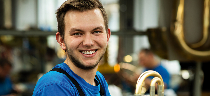 Eine junge Person in Arbeitskleidung sitzt lächelnd in einer Werkstatt, umgeben von Blasinstrumenten. Eine junge Person in Arbeitskleidung sitzt lächelnd in einer Werkstatt, umgeben von Blasinstrumenten.