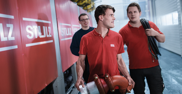 Drei junge Menschen gehen an einer Baustellenwand mit Firmenlogo entlang, einer davon trägt ein großes Rohrteil in der Hand Drei junge Menschen gehen an einer Baustellenwand mit Firmenlogo entlang, einer davon trägt ein großes Rohrteil in der Hand