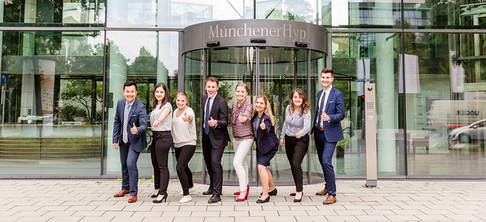 Gruppenbild von 8 jungen Menschen vor dem Eingang der Münchener Hypothekenbank Gruppenbild von 8 jungen Menschen vor dem Eingang der Münchener Hypothekenbank