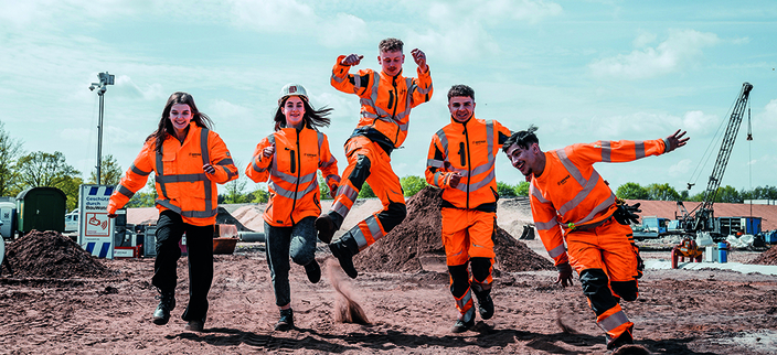 Fünf junge Personen in orangefarbener Arbeitskleidung laufen über eine Baustelle Fünf junge Personen in orangefarbener Arbeitskleidung laufen über eine Baustelle