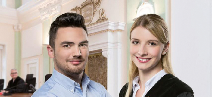 Zwei Personen lächeln in die Kamera im Hintergrund ist ein Gerichtssaal zu sehen. Zwei Personen lächeln in die Kamera im Hintergrund ist ein Gerichtssaal zu sehen.