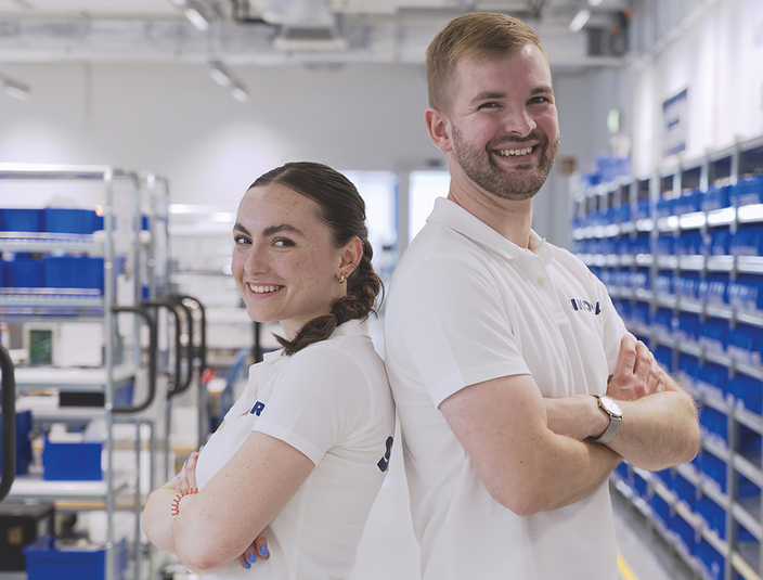 Zwei junge lächelnde Menschen in weißen Poloshirts mit Firmenlogo stehen Rücken an Rücken mit verschränkten Armen vor Regalen mit blauen Kästen Zwei junge lächelnde Menschen in weißen Poloshirts mit Firmenlogo stehen Rücken an Rücken mit verschränkten Armen vor Regalen mit blauen Kästen