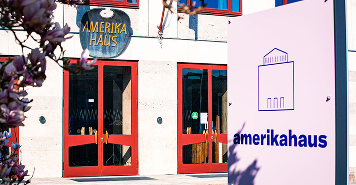 Ein schild mit amerikahaus Logo im Vordergrund. Im Hintergrund ein AHus mit 2 großen Glastüren mit rotem Rahmen