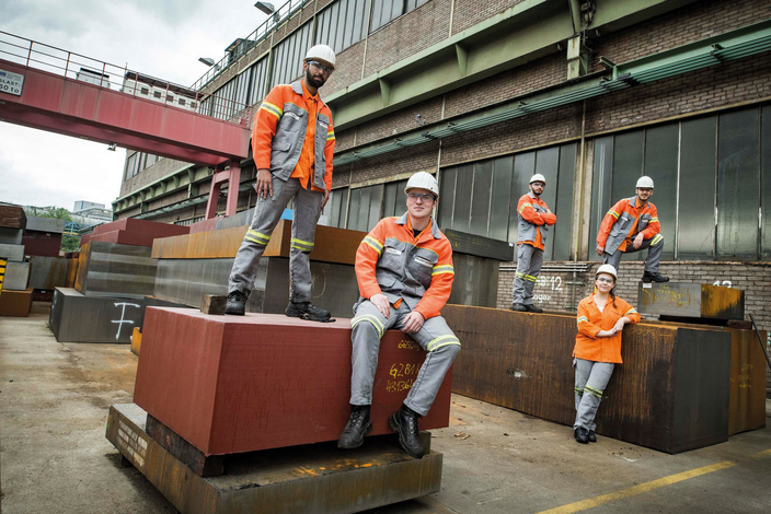 Fünf Personen in Schutzkleidung und Helmen posieren selbstbewusst in einem Industriehof zwischen großen Metallblöcken. Fünf Personen in Schutzkleidung und Helmen posieren selbstbewusst in einem Industriehof zwischen großen Metallblöcken.