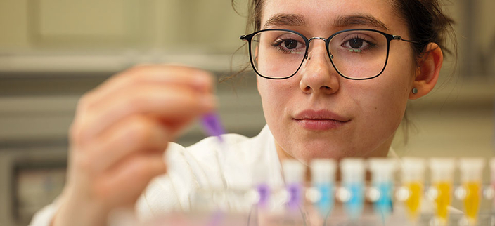 Eine Person im Laborkittel hält konzentriert eine Pipette über bunte Flüssigkeiten in kleinen Reagenzgläsern. Eine Person im Laborkittel hält konzentriert eine Pipette über bunte Flüssigkeiten in kleinen Reagenzgläsern.