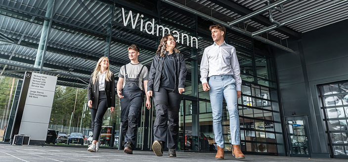 Vier junge Menschen in unterschiedlicher Berufskleidung gehen lächelnd aus einem Autohaus mit der Aufschrift "Widmann“. Vier junge Menschen in unterschiedlicher Berufskleidung gehen lächelnd aus einem Autohaus mit der Aufschrift "Widmann“.
