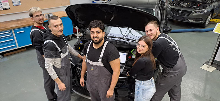 Fünf junge lächelnde Menschen in Arbeitskelidung stehen vor einem Auto mit geöffneter Motorhaube
