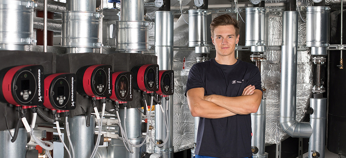 Junge Person mit verschränkten Armen vor einer Anlage für Gebäudetechnik