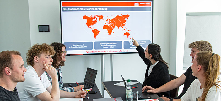 Sechs junge Personen sitzen an einem Tisch, auf dem davorstehenden Monitor eine Karte mit der Überschrift: Das Unternehmen: Marktbearbeitung Sechs junge Personen sitzen an einem Tisch, auf dem davorstehenden Monitor eine Karte mit der Überschrift: Das Unternehmen: Marktbearbeitung