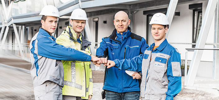 4 Mitarbeitende in Arbeitskleidung im Aussengelände die ihre Fäuste aneinanderhalten 4 Mitarbeitende in Arbeitskleidung im Aussengelände die ihre Fäuste aneinanderhalten