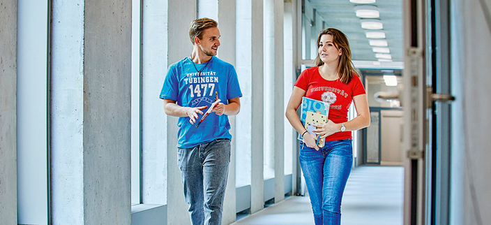 Zwei junge Menschen in T-Shirts der Universität Tübingen gehen lächelnd durch einen hellen Flur und unterhalten sich. Zwei junge Menschen in T-Shirts der Universität Tübingen gehen lächelnd durch einen hellen Flur und unterhalten sich.