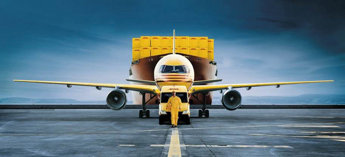 Person in gelber Arbeitskleidung vor einem Transporter, einem Flugzeug und einem Containerschiff
