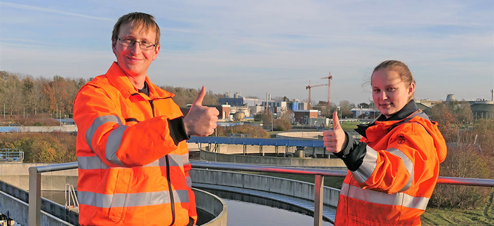 Zwei Personen in orangefarbener Warnkleidung stehen vor einer Kläranlage und zeigen beide lächelnd den Daumen nach oben.