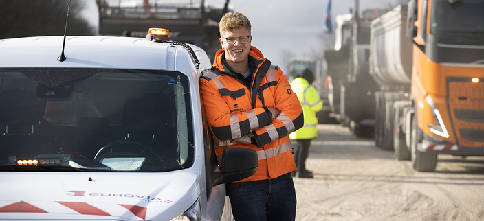 Junge lächelnde Person in orangefarbener Jacke lehnt an einem EUROVIA-Wagen, im Hintergrund Baustellenfahrzeuge
