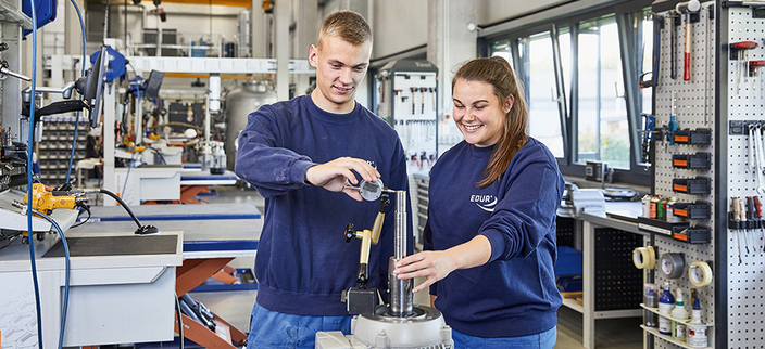 Zwei junge lächelnde Mitarbeitende arbeiten an einer Vorrichtung in der Produktionsstätte Zwei junge lächelnde Mitarbeitende arbeiten an einer Vorrichtung in der Produktionsstätte