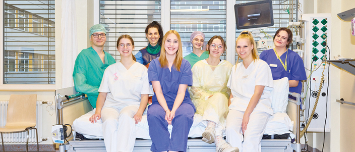 Mitarbeitende in ihrer jeweiligen Arbeitskleidung aus den verschiedenen Bereichen der Helios Klinik auf einem Krankenbett versammelt Mitarbeitende in ihrer jeweiligen Arbeitskleidung aus den verschiedenen Bereichen der Helios Klinik auf einem Krankenbett versammelt