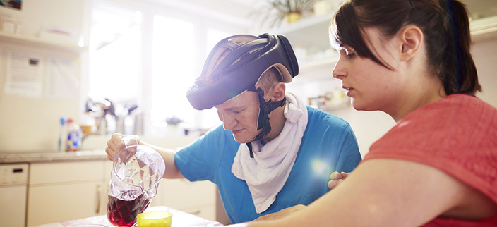 Junge Person betreut eine ältere Person mit Helm, die sich etwas zu trinken eingießt Junge Person betreut eine ältere Person mit Helm, die sich etwas zu trinken eingießt