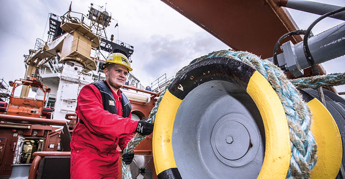 Person in roter Arbeitskleidung mit Helm zieht an einem dicken Tau, Containerschiff im Hintergrund