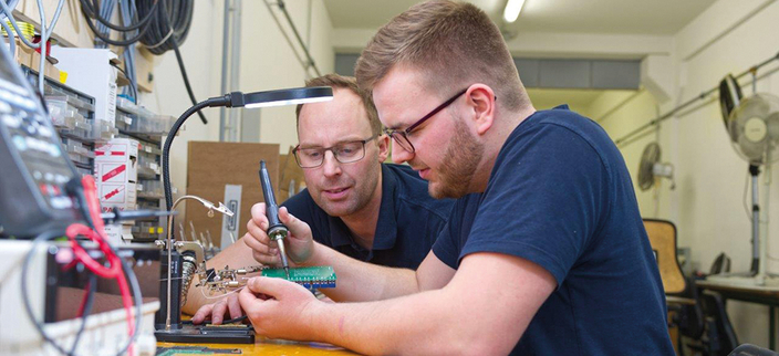 Zwei Menschen in blauen T-Shirts mit Brille arbeiten an einer Platine