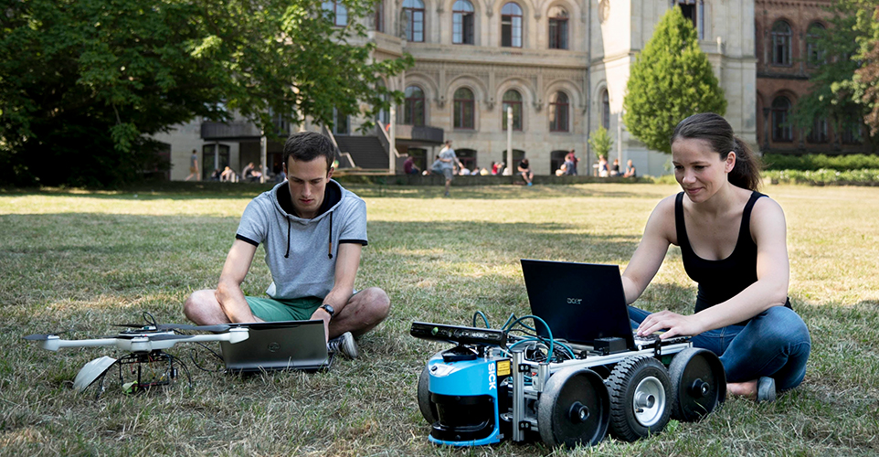 2 Menschen sitzen vor Unigebäude auf der Wiese mit Laptops und kleinen technischen Maschinen
