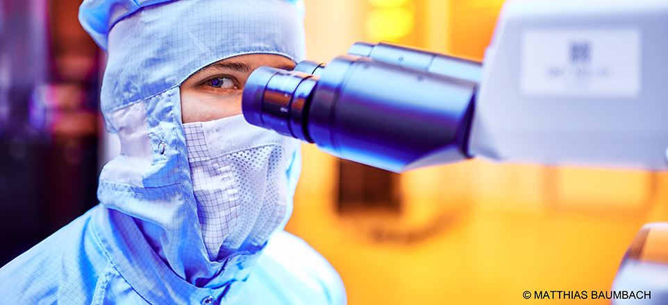Person in Schutzkleidung sitzt an einem Mikroskop im Labor