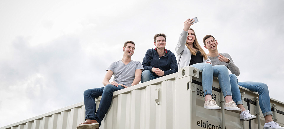 Vier junge lächelnde Personen sitzen auf einem Container und machen ein Selfie