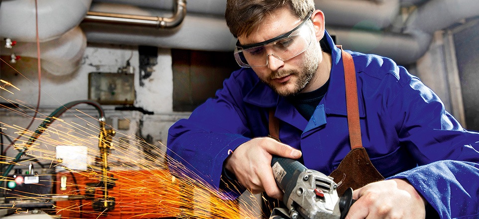 Person in blauer Arbeitskleidung mit Schutzbrille arbeitet mit einem Gerät, Funken sprühen