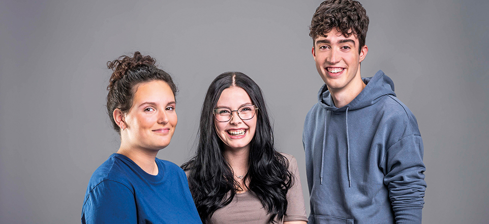 Drei junge Personen vor einem grauen Hintergrund lächeln in die Kamera