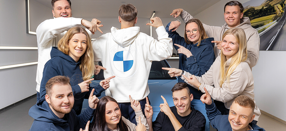 Gruppenfoto von neun jungen lächelnden Personen, die auf den Rücken einer Person im Kapuzenpulli it BMW-Logo zeigen