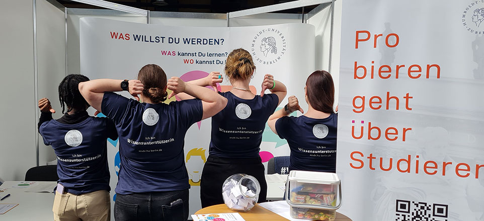 Mitarbeitende der Humboldt Universität zu Berlin am Messestand zeigen mit den Daumen auf das Logo auf dem Rücken der T-Shirts