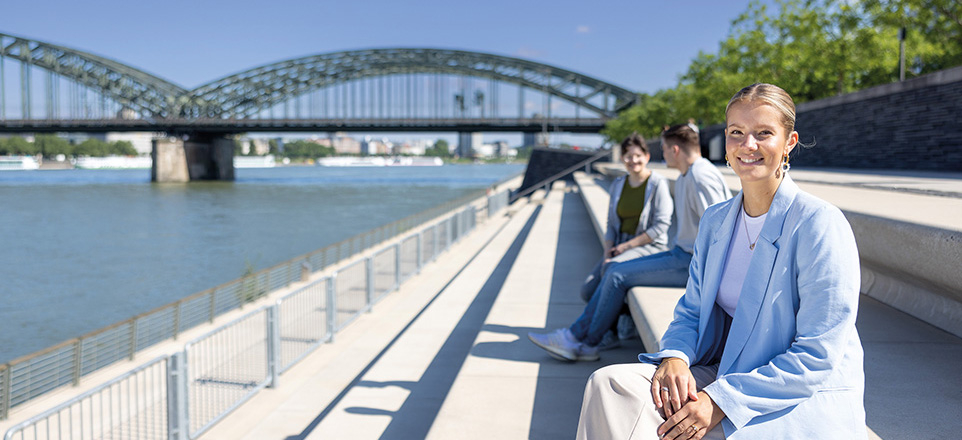 Junge Frau sitzt auf einer Steintreppe am Rhein und lächelt in die Kamera