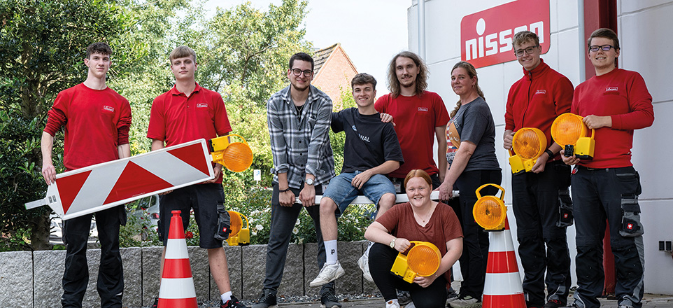 Teamfoto der Auszubildenden mit Baustellen-Warnleuchten und Verkehrskegeln vor dem Firmengebäude