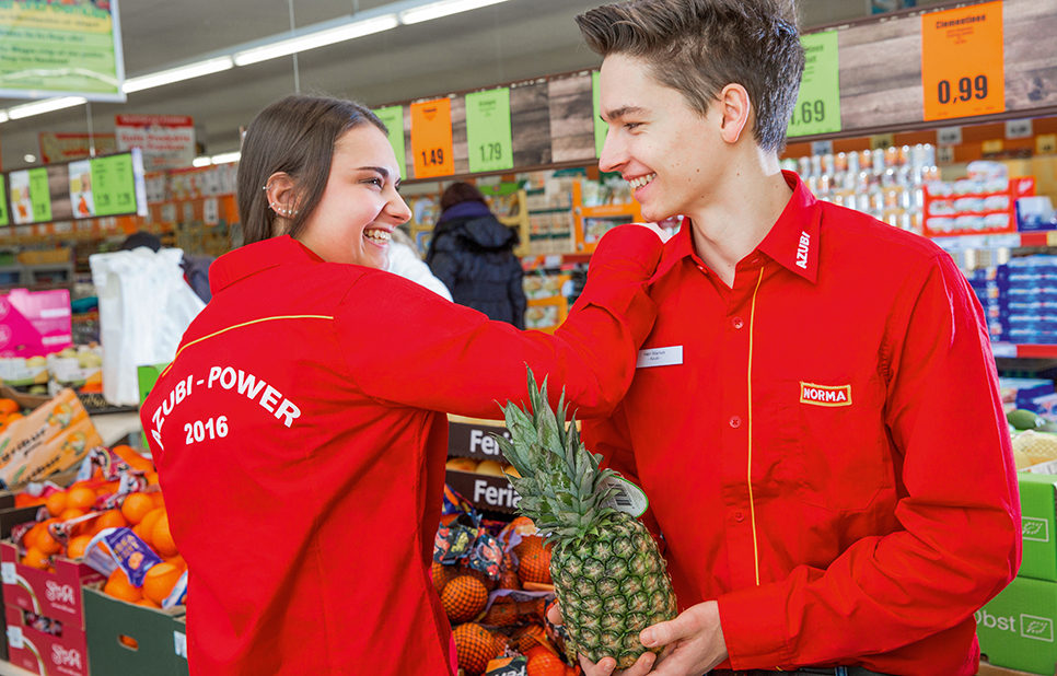 Zwei junge Menschen in roten Hemden mit Firmenlogo und der Aufschrift Azubi-Power in der NORMA-Filiale, einer davon hat eine Ananas in der Hand