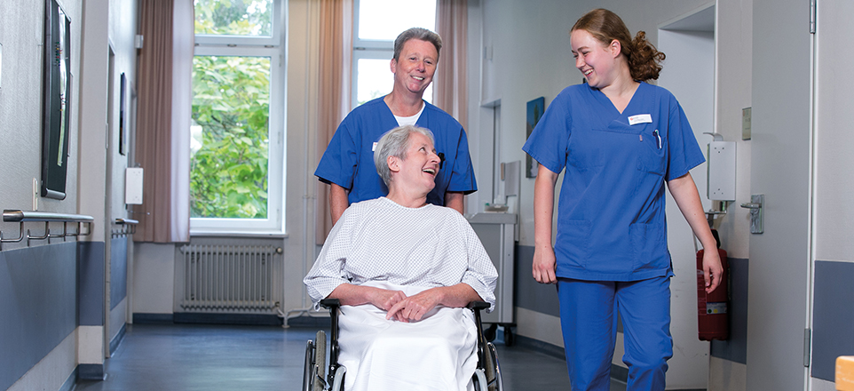 Zwei Krankenschwestern schieben Frau im Rollstuhl durch Krankenhausflur Zwei Krankenschwestern schieben Frau im Rollstuhl durch Krankenhausflur