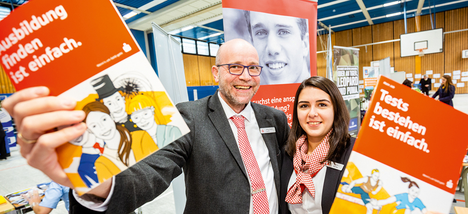 Zwei Mitarbeitende der Sparkasse halten lachend Ausbildungsflyer in die Kamera